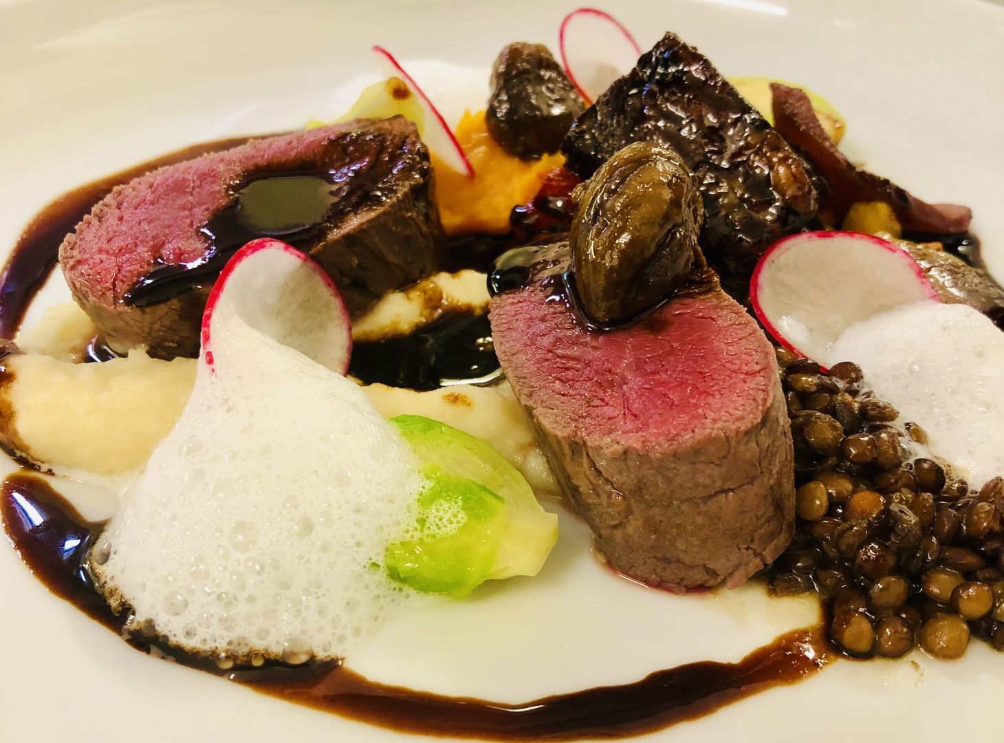 Zwei rosa gebratene Fleischstücke auf weißem Teller mit dunkler Sauce, Schaum, Linsen, Pilzen und Radieschenscheiben angerichtet.